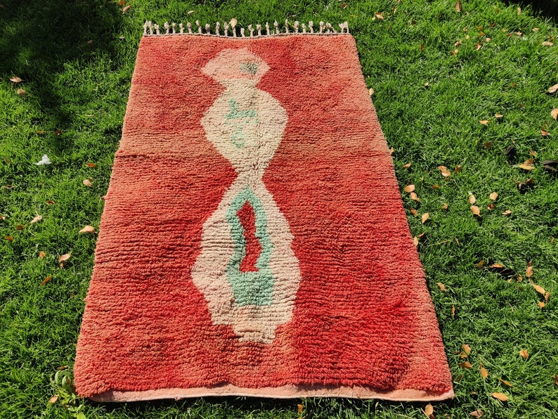Puede incluir: Una alfombra roja marroqu&iacute; con un patr&oacute;n geom&eacute;trico blanco y verde. La alfombra tiene un borde de flecos.