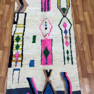 Peut inclure: Un tapis blanc avec un motif g&eacute;om&eacute;trique dans des tons de rose, bleu, vert, jaune, noir et gris. Le tapis a une bordure frang&eacute;e.