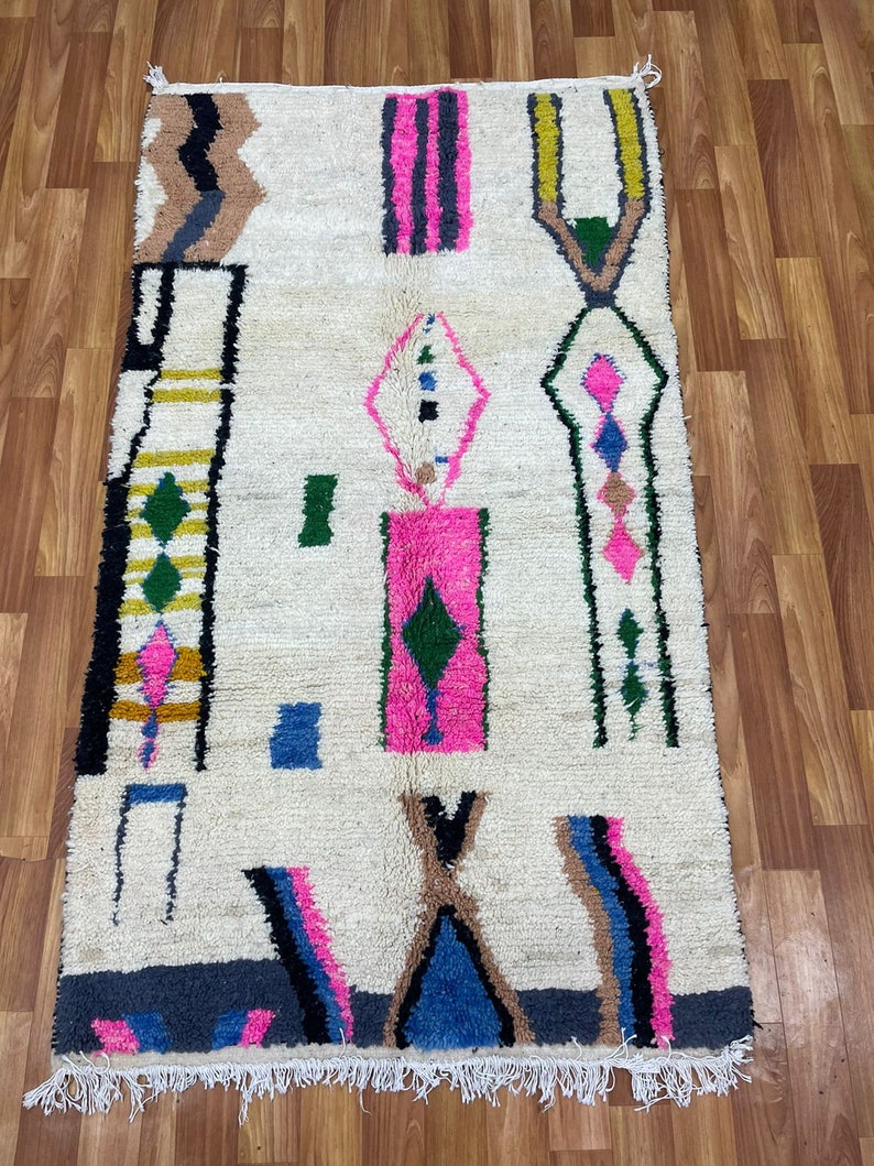 Peut inclure: Un tapis blanc avec un motif g&eacute;om&eacute;trique dans des tons de rose, bleu, vert, jaune, noir et gris. Le tapis a une bordure frang&eacute;e.