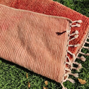 Puede incluir: Dos alfombras tejidas de color rosa y rojo con flecos. Las alfombras est&aacute;n dispuestas sobre un lecho de hierba verde.