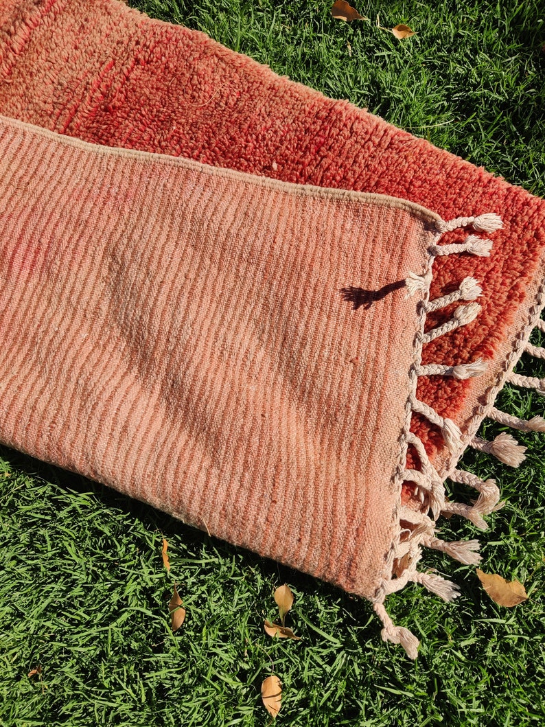 Puede incluir: Dos alfombras tejidas de color rosa y rojo con flecos. Las alfombras est&aacute;n dispuestas sobre un lecho de hierba verde.