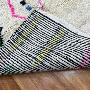 Peut inclure: Un tapis blanc et moelleux avec un motif g&eacute;om&eacute;trique color&eacute;. Le tapis a des accents de rose, bleu, jaune, noir et vert. Le tapis est pli&eacute; en deux, montrant le dessous du tapis, qui est un tissu blanc avec des rayures noires.