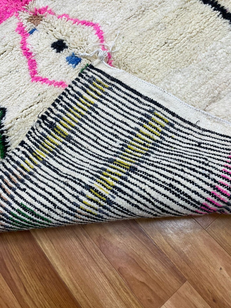 Peut inclure: Un tapis blanc et moelleux avec un motif g&eacute;om&eacute;trique color&eacute;. Le tapis a des accents de rose, bleu, jaune, noir et vert. Le tapis est pli&eacute; en deux, montrant le dessous du tapis, qui est un tissu blanc avec des rayures noires.