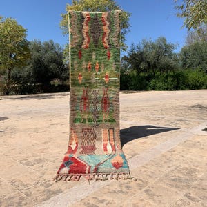Puede incluir: Una alfombra rectangular, de orientación vertical, con un diseño geométrico colorido. La alfombra presenta un fondo verde con detalles rojos, naranjas y azules. Tiene un borde con flecos y se exhibe al aire libre.