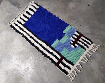 Puede incluir: Una alfombra colorida con un borde rayado en blanco y negro y un patrón geométrico azul, verde y azul claro en el centro.