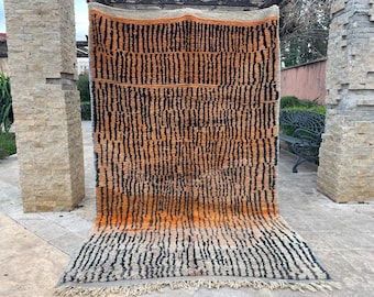 Puede incluir: Una alfombra marroquí tejida a mano con un patrón geométrico en tonos naranja, negro y blanco. La alfombra tiene un borde de flecos y está hecha de lana.
