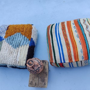 K&ouml;nnte beinhalten: Zwei bunte, gemusterte Bodenkissen, eines mit einem geometrischen Muster in Blaut&ouml;nen, Wei&szlig; und Orange, das andere mit einem Streifenmuster in Orange-, Blau- und Wei&szlig;t&ouml;nen. Ein kleiner Holzhocker mit einem braunen Tontopf steht vor den Kissen.
