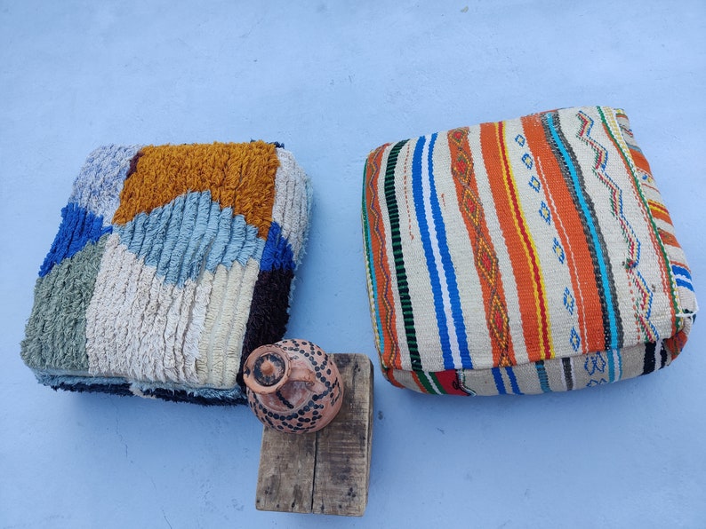 K&ouml;nnte beinhalten: Zwei bunte, gemusterte Bodenkissen, eines mit einem geometrischen Muster in Blaut&ouml;nen, Wei&szlig; und Orange, das andere mit einem Streifenmuster in Orange-, Blau- und Wei&szlig;t&ouml;nen. Ein kleiner Holzhocker mit einem braunen Tontopf steht vor den Kissen.