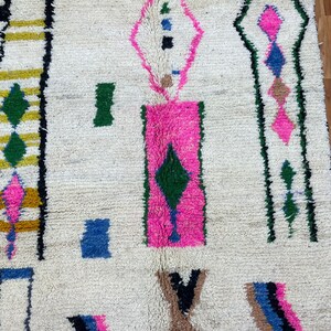 Peut inclure: Un tapis marocain blanc avec un motif g&eacute;om&eacute;trique en rose, vert, bleu et noir. Le tapis a une texture moelleuse et est fait de laine.