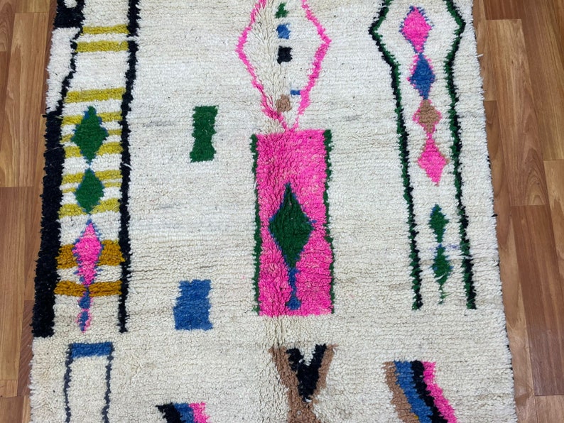 Peut inclure: Un tapis marocain blanc avec un motif g&eacute;om&eacute;trique en rose, vert, bleu et noir. Le tapis a une texture moelleuse et est fait de laine.