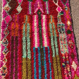 Puede incluir: Una alfombra marroqu&iacute; colorida con un patr&oacute;n geom&eacute;trico en tonos de rosa, rojo, verde, azul y marr&oacute;n. La alfombra tiene un borde de flecos y est&aacute; hecha de lana.