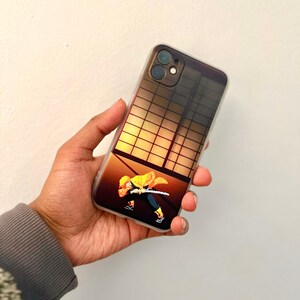 May include: Clear phone case with a vibrant anime character design. The case shows a figure with a sword against a sunset backdrop with a grid pattern. The phone case is held in a hand.