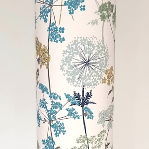 May include: A cylindrical lampshade featuring a floral design in teal, olive green, and gold on a white background. The lamp has a white metal base with three legs. The height is approximately 100 cm.