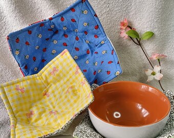 May include: Three fabric bowl cosies featuring distinct patterns: one blue with ladybugs and white flowers, one yellow with a gingham check and pink bows, and one green with a small floral print, holding an orange and white ceramic bowl.