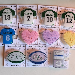 Może przedstawiać: Kolekcja różnych przedmiotów dekoracyjnych, w tym wzory w kształcie koszulki i serca, a także piłka do rugby i iPoda. Koszulki mają imiona i numery, serca inicjały. Ekran iPoda wyświetla listę piosenek.