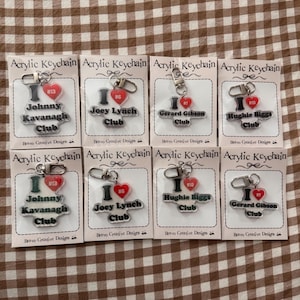 May include: Acrylic keyrings with the text "I <3" and names such as "Johnny Kavanagh Club" and "Joey Lynch Club." Each keyring has a heart charm and a silver-coloured keyring. The keyrings are displayed on small white cards.