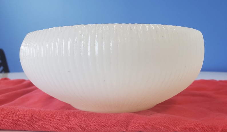 May include: A white, ribbed glass bowl with a rounded shape. The bowl is sitting on a red fabric surface.