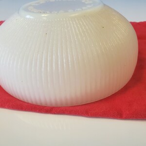 May include: A white, ribbed, ceramic bowl with a scalloped rim. The bowl is sitting on a red cloth.