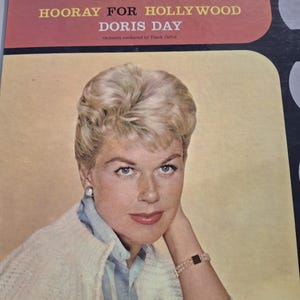 May include: Vintage vinyl record album cover for "Hooray for Hollywood" by Doris Day. The cover features a portrait of Doris Day with blonde hair, wearing a light blue striped shirt and a white cardigan. The album title and artist name are in red and gold.