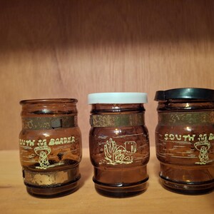 May include: Three brown glass jars with cork lids. Each jar has a white label with the text "South of the Border" and a cartoon image. The jars are on a wooden surface.