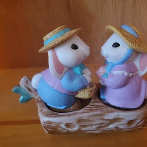 May include: A pair of ceramic bunny salt and pepper shakers. The bunnies are wearing straw hats and holding baskets. They are sitting on a brown log base.