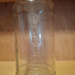 May include: A clear glass bottle with a metal cap. The bottle has embossed text and measurement markings. The bottle is cylindrical with a slightly rounded base and a narrow neck. The background is a wooden surface.
