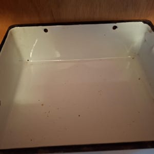 May include: A rectangular, white enamelware container with a black rim. The interior is white with minor imperfections. The container has three small holes on the sides. The container is empty.