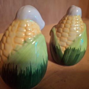 May include: Two ceramic salt and pepper shakers shaped like corn on the cob. The shakers are painted to look like corn on the cob with green husks and yellow kernels. The tops of the shakers are white.