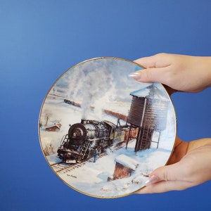 May include: A vintage-style porcelain plate with a gold rim features a winter scene with a steam engine train travelling through a snowy landscape. The train is black with gold accents and is pulling a string of boxcars. The scene also includes a water tower and a small building.