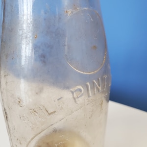 May include: A clear glass bottle with the embossed text "HALF PINT" and a circular design. The bottle has a curved shape and appears vintage, with some wear and discoloration. The background is a gradient of white and blue.