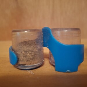 May include: Two small, clear plastic containers with a blue plastic holder. One container is filled with a granular substance. The containers are on a wooden surface.