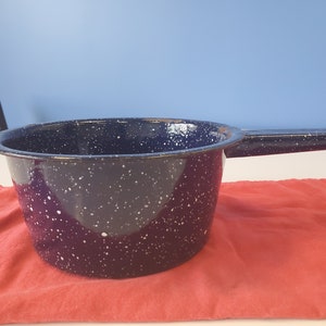 May include: A navy blue enamel saucepan with a long handle and white speckles. The pot sits on a red cloth. The kitchenware is designed for cooking and is suitable for various culinary tasks.