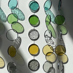 Op de afbeelding: Een mobiel gemaakt van ronde glazen stukken in verschillende kleuren en texturen, waaronder groen, geel en grijs. De glazen stukken zijn opgehangen aan zilveren kettingen.