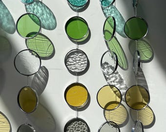 May include: A mobile made of round glass pieces in various colours and textures, including green, yellow, and grey. The glass pieces are suspended from silver chains.
