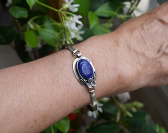 May include: Silver bracelet featuring an oval, deep blue gemstone. The bracelet has a floral design on the sides of the gemstone and is worn on a person's wrist. The background includes green foliage and white flowers.