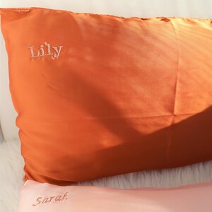 Puede incluir: Dos fundas de almohada de sat&eacute;n, una naranja y otra rosa, con los nombres "Lilly" y "Sarah" bordados.