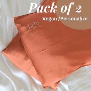Puede incluir: Dos fundas de almohada de color &oacute;xido con la palabra "Lily" bordada en una de ellas. Las fundas de almohada est&aacute;n hechas de materiales veganos y se pueden personalizar. El texto "Pack of 2 Vegan/Personalize" es visible en la imagen.