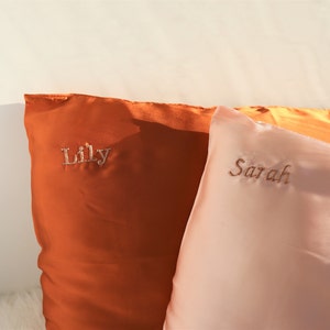 Puede incluir: Dos fundas de almohada de sat&eacute;n, una naranja y otra rosa, con los nombres "Lily" y "Sarah" bordados en cursiva.