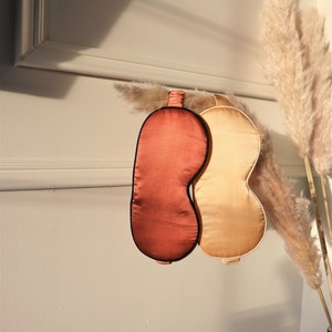 Puede incluir: Dos máscaras para dormir, una es de color rojo oscuro y la otra es de color beige claro. Ambas máscaras tienen un borde negro alrededor de los bordes.