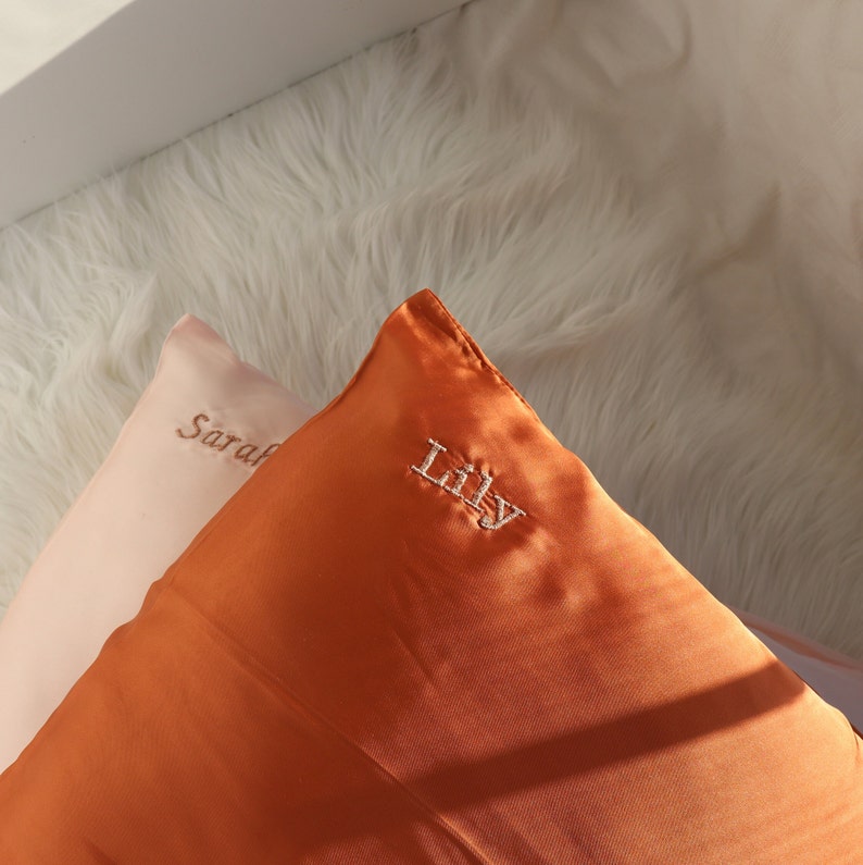Puede incluir: Dos fundas de almohada de sat&eacute;n, una melocot&oacute;n y otra naranja, con los nombres "Sarah" y "Lilly" bordados.