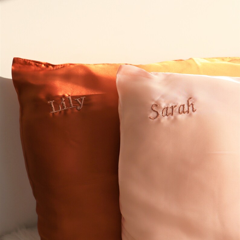 Puede incluir: Dos fundas de almohada de sat&eacute;n con nombres bordados, "Lily" en una funda de almohada naranja y "Sarah" en una funda de almohada melocot&oacute;n.
