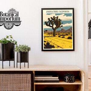 May include: A framed poster with a vintage illustration of a Joshua Tree in the desert. The poster is titled "Joshua Tree, California". The poster is hanging on a white wall above a wooden shelf with black metal plant stands and potted plants.
