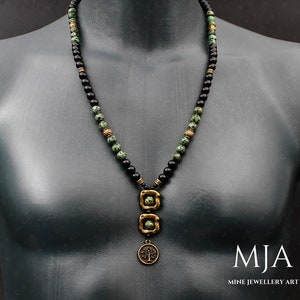 May include: A black and green beaded necklace with a gold pendant featuring a tree design. The pendant is made of two square shapes with a circle attached to the bottom. The necklace is on a mannequin.