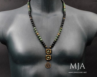 May include: A black and green beaded necklace with a gold pendant featuring a tree design. The pendant is made of two square shapes with a circle attached to the bottom. The necklace is on a mannequin.