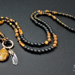 May include: A layered necklace featuring black and brown beads, a gold pendant with a tree design, and two silver feathers. The necklace is displayed against a dark backdrop.