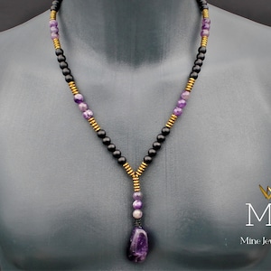May include: A necklace with black and gold beads and a large amethyst pendant. The necklace is on a mannequin. The text "MJA Mine Jewellery Art" is in the bottom right corner of the image.