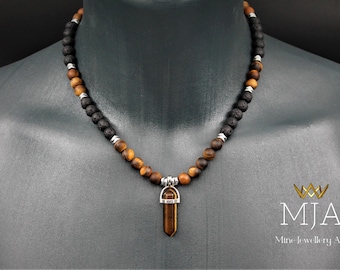 Pode incluir: Um colar com contas marrons e pretas e um pingente prateado com uma pedra de olho de tigre. O colar está em um manequim preto. O texto "MJA Mine Jewellery Art" está no canto inferior direito da imagem.