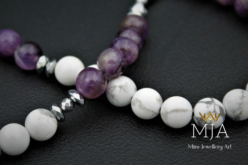 K&ouml;nnte beinhalten: Nahaufnahme eines Armbands aus wei&szlig;en und violetten Perlen. Das Armband besteht aus wei&szlig;en Howlith-Perlen, violetten Amethyst-Perlen und silbernen Metallperlen. Das Armband liegt auf einer schwarzen Oberfl&auml;che. Der Text "MJA Mine Jewellery Art" ist in der unteren rechten Ecke des Bildes sichtbar.