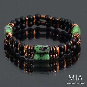May include: A double-strand bracelet with black onyx beads, copper spacers, and a green and pink gemstone focal bead. The bracelet is on a gray background. MJA Mine Jewellery Art.