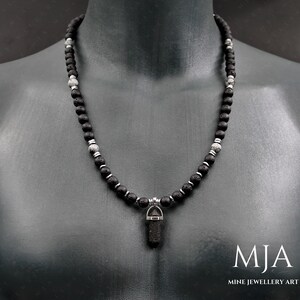 May include: A black and silver beaded necklace with a black lava stone pendant. The necklace is on a mannequin's neck. The text "MJA MINE JEWELLERY ART" is at the bottom of the image.